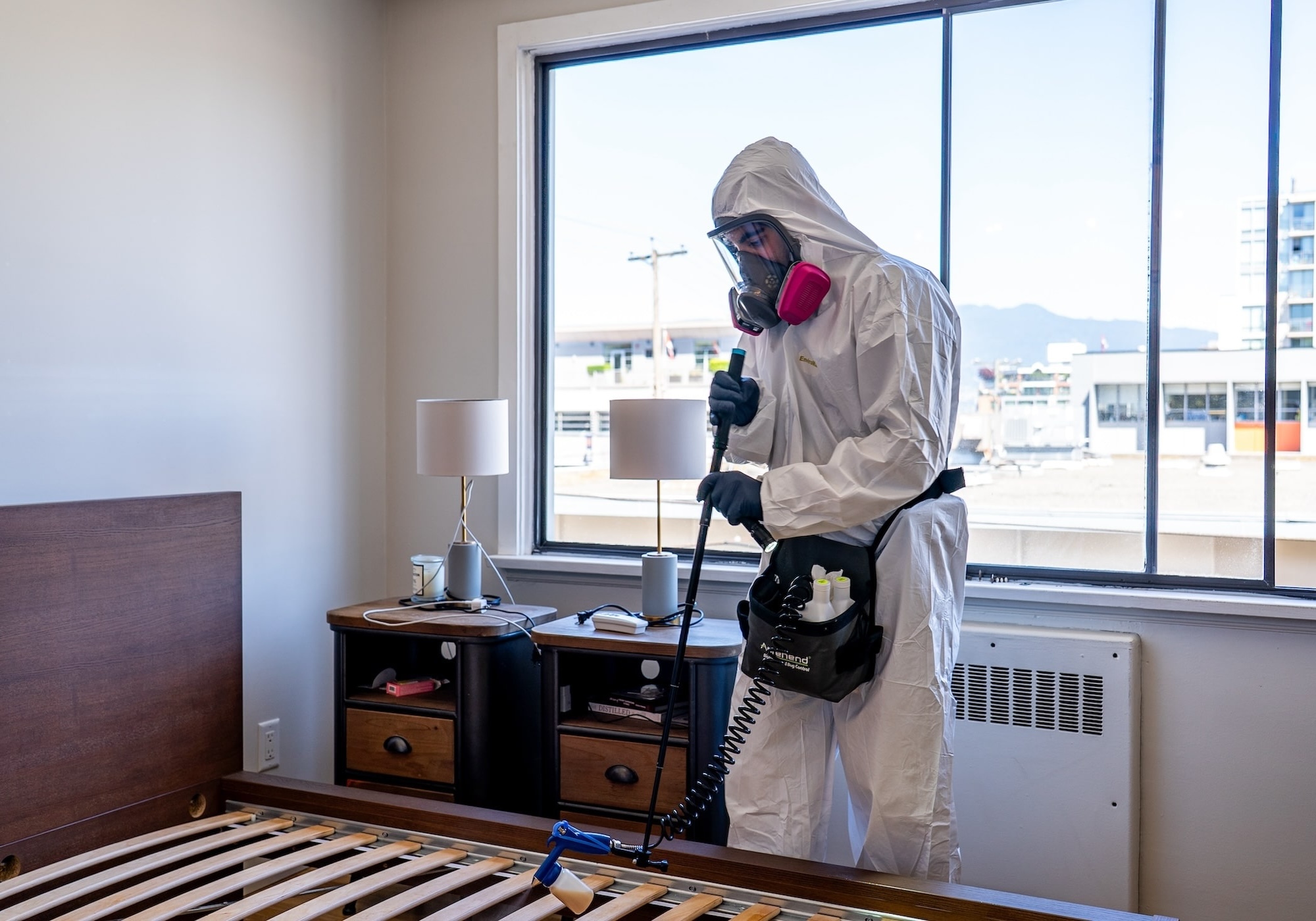 Phantom Pest Control technician in a protective suit treating a bed frame in a condo bedroom