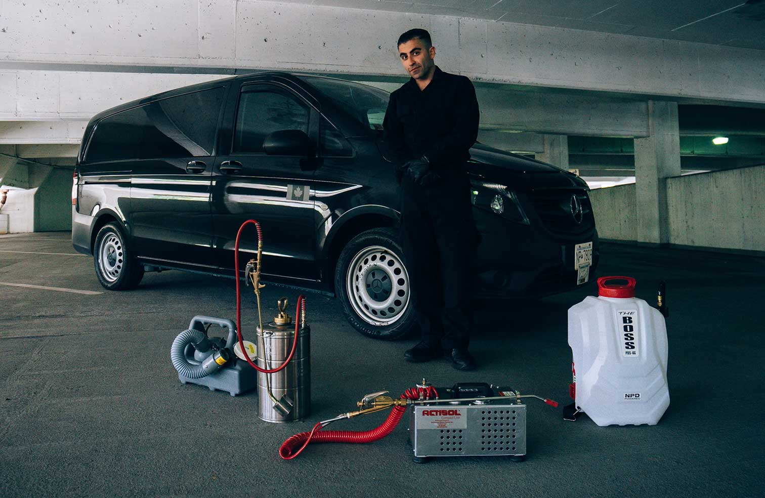 A Phantom Pest Control technician standing beside a service van in a parkade, with spray equipment and a sprayer on the ground